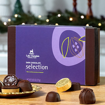 Dark chocolate 15 piece assortment sitting on a wooden table with assorted wrapped and unwrapped chocolates around the box and greenery with Christmas lights on it in the background.