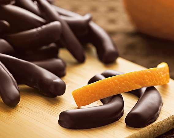 Pile of dark chocolate covered orange peels with an orange peel laying over two dark chocolate covered orange peels, on a wooden cutting board.
