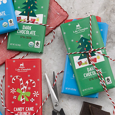 Assorted milk and dark chocolate bars wrapped in holiday themed packaging and a white and red stripped twine wrapped around a pike of chocolate bars and a pair of scissors trying to cut the twine.