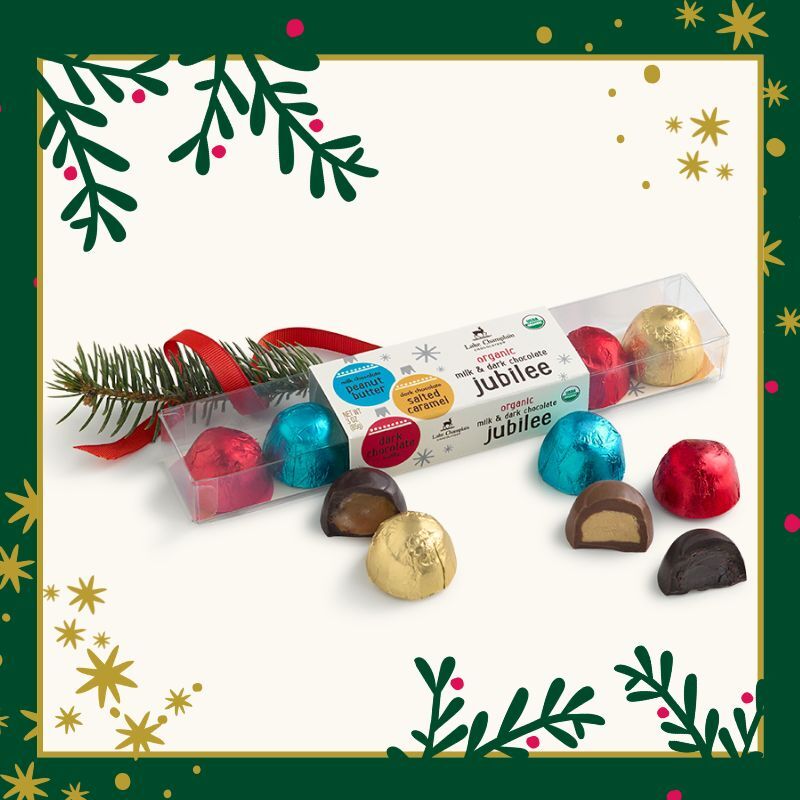 Clear assorted jubilee gift box with wrapped and unwrapped jubilees around the box and a Christmas tree branch with a red ribbon wrapped around it and Christmas themed art around the gift box.