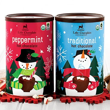 Peppermint hot chocolate and Winter traditional hot chocolate  sitting on a white wooden table with red snowflakes under the tins and pieces of peppermint around the Peppermint can and pieces of chocolate around the Winter can.