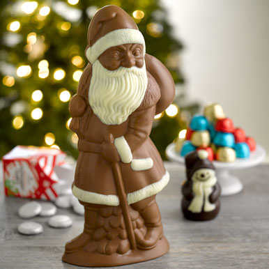 Giant milk chocolate Santa with white chocolate accents sitting on a gray wooden counter with holiday chocolates and a Christmas tree with white lights in the background.