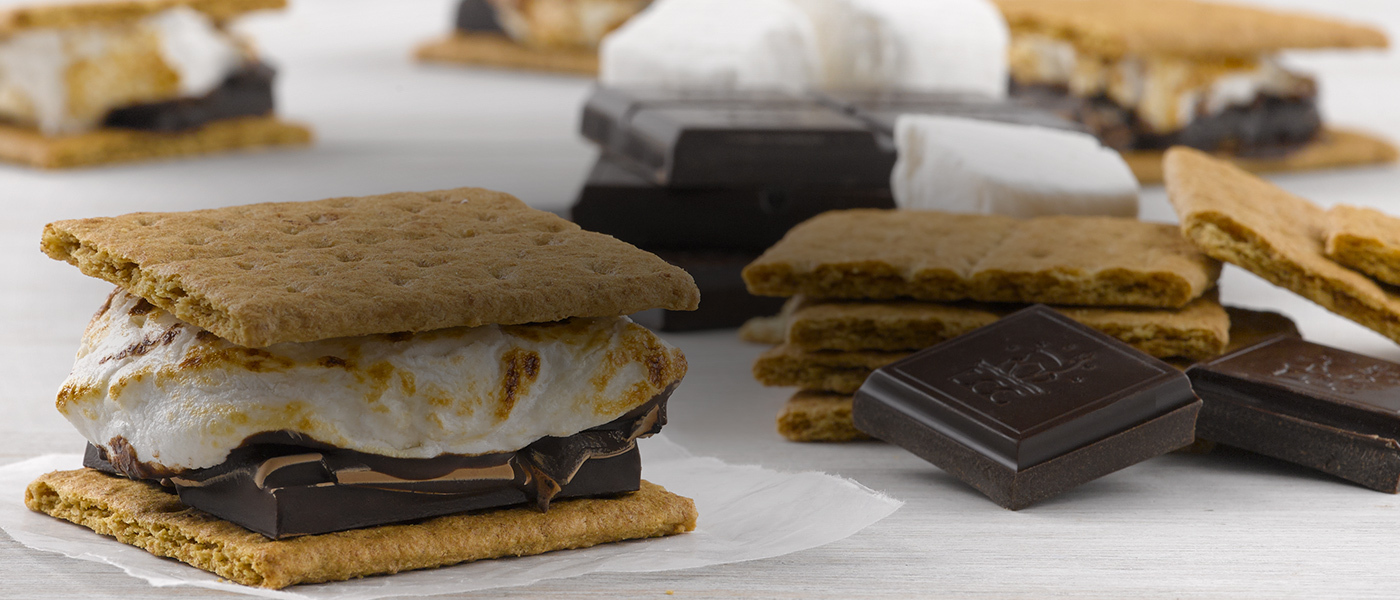 Close up image of gourmet s'mores made with graham crackers, melted chocolate bars, and roasted marshmallows, sitting on a white wood surface.