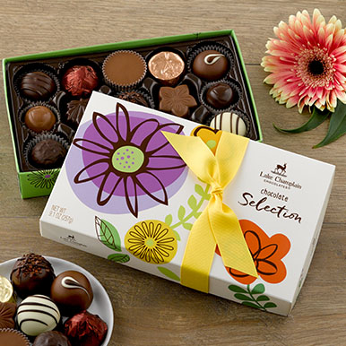 Assorted gourmet chocolates in a spring themed gift box with a yellow ribbon and a white ceramic circular dish with assorted chocolates on it and a pink flower in the corner next to the gift box.