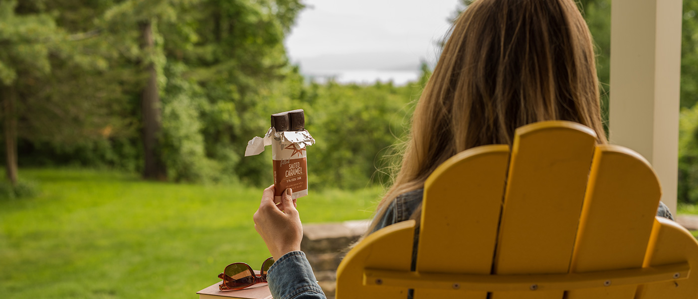 Woman sitting outside in an Adirondack chair holding a partially opened dark chocolate salted caramel bar in her left hand while looking at a view of trees and the lake.