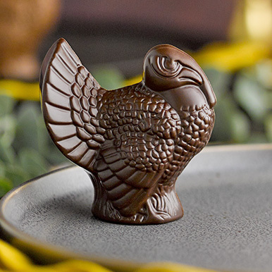 Dark chocolate turkey favor sitting on a gray stone plate with greenery, gold candlesticks and napkins in the background.