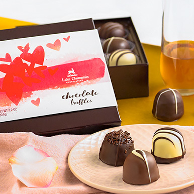 Assorted chocolate truffles on a white ceramic plate with a Valentine's Day themed gift box in the background and a glass of champagne.