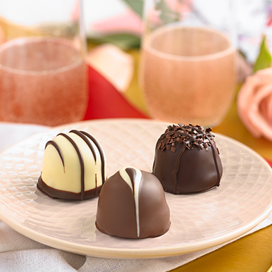A white chocolate raspberry truffle, cappuccino truffle and french roast truffle sitting on a white ceramic plate sitting on a gold tray with two glasses of sparkling pink champagne in the background