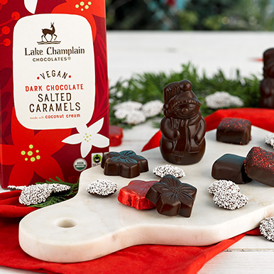 Assorted dark vegan holiday chocolates sitting on a marble cutting board on top of a red napkin with holiday greenery in the background.