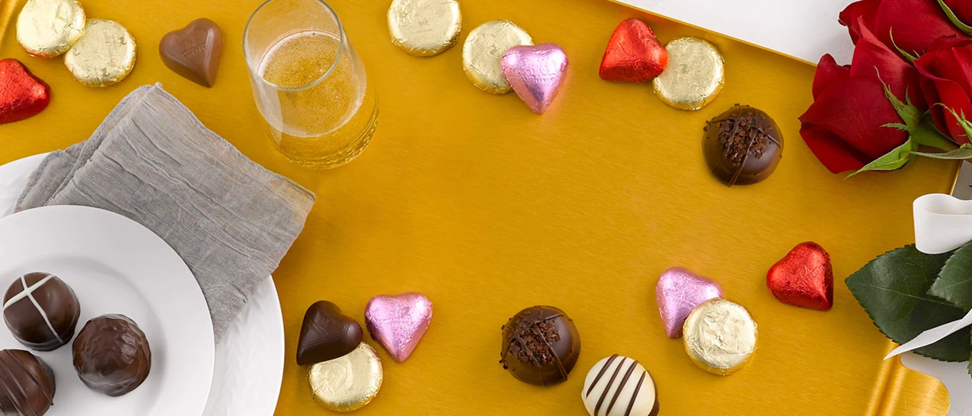Assorted milk and dark chocolate coins, hearts, and gourmet truffles sitting on a gold tray with a glass of champagne and red roses and a white round plate with gourmet truffles on it with a gray napkin underneath. 