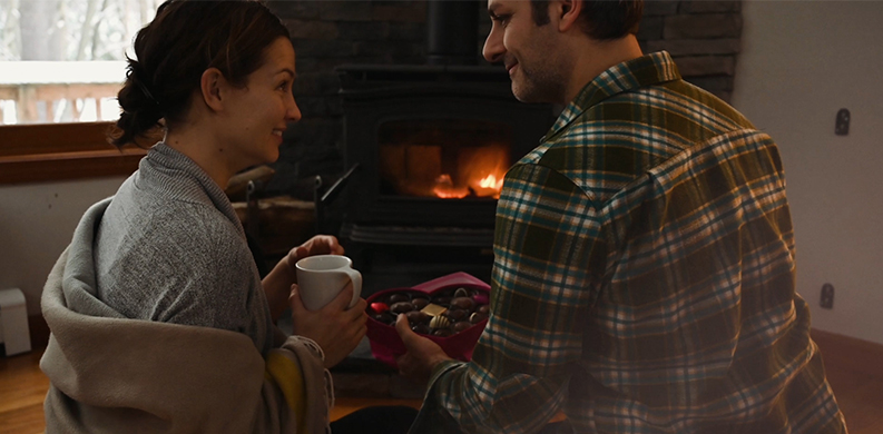 couple with a heart-shaped box of chocolate in front of a fireplace