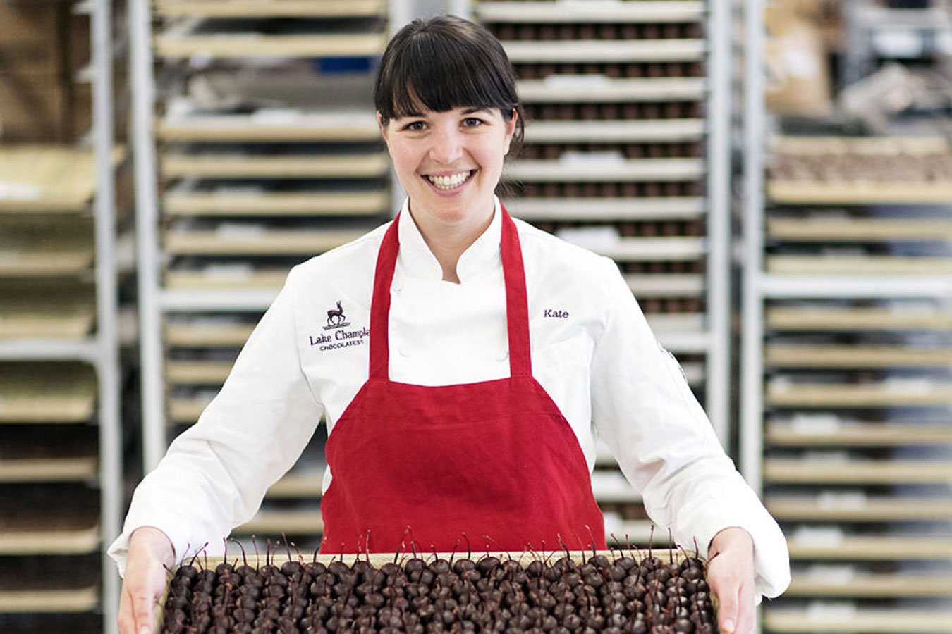 Woman wearing a white shirt with LCC logo and a red apron, holding a tray of chocolate covered cherries.