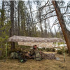 Outdoor picture of the SJK Satellite Tarp setup in the woods with a hunter unpacking his backpack underneath it.