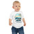 Front view of baby model wearing white baby t-shirt with graphic design of animated sunrise on tropical island, navy blue text 'Hakuna Aloha It means No Laundry!