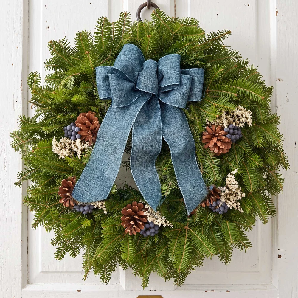 An elegant Blueberry fresh Maine balsam wreath with a blue linen bow and white statice pops displayed on a white wooden door.