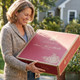 A smiling woman holding Harbor Farm's signature red gift box, ready to deliver a forest-fresh Maine wreath.