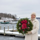 Woman holding a Winterberry fresh Maine balsam Christmas wreath beside a snowy coastal harbor