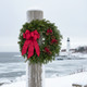 A 22-inch Cape Elizabeth fresh balsam wreath displayed on a post overlooking a snowy coastal Maine lighthouse during a winter storm.