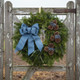 A fresh Bar Harbor balsam wreath with blue ribbon hanging on a rustic wire and wood farm gate.