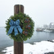A Bar Harbor fresh balsam wreath with a blue bow mounted on a wooden post in a foggy Maine coastal harbor with fishing boats.
