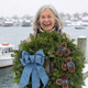 Close-up of a smiling woman holding a Bar Harbor balsam wreath with blue linen ribbon in a snowy Maine fishing village setting.