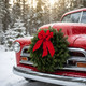 A Classic Maine balsam wreath with a red velvet bow mounted on the hood of a vintage red truck in a snowy evergreen forest.