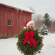 A happy young girl in a Santa hat holding a large, fresh balsam Christmas wreath in front of a snow-covered red barn in Maine.
