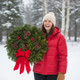 A laughing woman in a red jacket holding a Downeaster fresh balsam wreath in a snowy Maine forest, highlighting the wreath's large size and fullness.