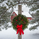 A rustic Downeaster fresh balsam wreath hanging on a wooden post with a snow-covered red barn and evergreen trees in the distance.