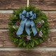 A lush Blueberry fresh Maine balsam wreath with a blue linen bow and white German statice hanging on a rustic log cabin wall.