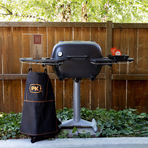 PK Grills PK360 Start Cookin' Bundle - Graphite, a portable charcoal grill with a black lid, silver stand, and side shelves, outdoor setting with a wooden fence and green foliage in the background.