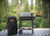 PK Grills PK300 Start Cookin' Bundle - Graphite, outdoor portable grill setup with cover and accessories on a wooden deck with a lush green forest background.