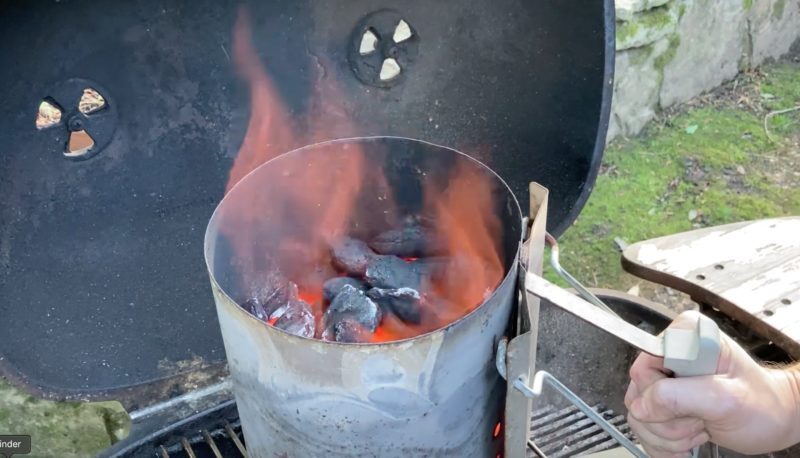 Chimney starter filled with burning charcoal briquettes producing orange flames inside an open PK grill, showing the ignition process before cooking.