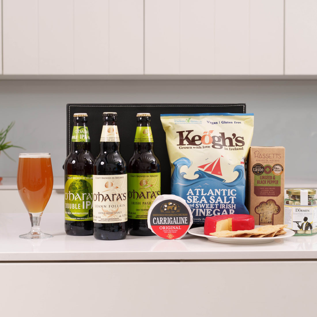 Big Night In Craft Beer Box. A selection of food and beverages is displayed on a countertop. Three bottles of beer labeled "O'Hara's" are arranged alongside a pint glass filled with amber liquid. Next to the beers, a bag of Keogh's Atlantic Sea Salt and Vinegar chips is visible. A round container of Carrigaline cheese sits beside a package labeled "Hassetts" and some crackers on a plate.