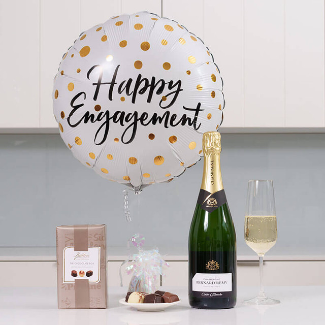Happy Engagement Balloon & Bubbles Gift. A white and gold polka dot balloon displays the text "Happy Engagement." Beside it, a bottle of champagne stands next to a glass filled with champagne. In the foreground, there is a box of chocolates and a small bag of treats. The setting features a modern kitchen background.