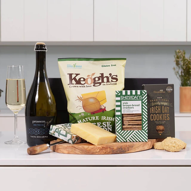 The Sparkling Picnic Treat Box. A display of gourmet food items arranged on a wooden platter. The arrangement includes a bottle of sparkling wine, a bag of Keogh's Irish cheese & onion crisps, a block of cheese, a box of chocolate biscuit bars, and a package of Irish oat cookies. A glass filled with sparkling wine is positioned nearby.