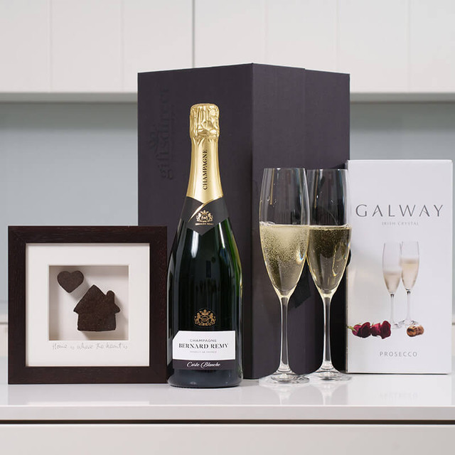 New Home Champagne Gift Set`. A bottle of champagne is placed next to two filled flutes on a countertop. A framed picture featuring a heart and house design is positioned in front of the champagne. Beside the frame, there is a box labeled "GALWAY CRYSTAL," showcasing additional glasses and chocolates.