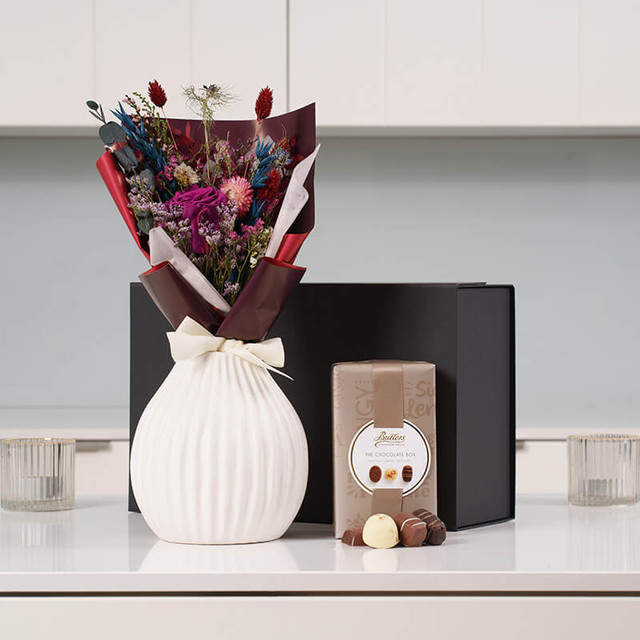 Mothers Day Plum Bouquet & Chocolates Gift. A white textured vase (not included) holds a bouquet of dried flowers in shades of pink, red, and blue. Next to the vase is a box of chocolates with a circular label on the front, placed on a light-colored countertop. A black gift box sits in the background.