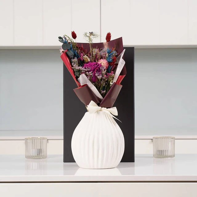 Plum Everblooms Bouquet Gift – vibrant floral arrangement in purple, red, and blue hues, wrapped in red and brown paper with white ribbon, presented in a ribbed white vase.