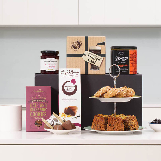 Mothers Day Afternoon Tea Treat Box: A decorative display of assorted treats on a white countertop. Included are a two-tier serving stand with baked goods, a box of chocolate truffles, jars of jam and beverage mix, and a package of cookies.