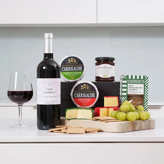 Red Wine & Cheese Gift Box. A wooden serving board displays various cheeses, crackers, and green grapes. In the background, there are two rounds of Carrigaline cheese and a jar of chutney. A bottle of red wine and a glass filled with wine are placed beside the board. The setting features a light-colored countertop against a soft blue wall.