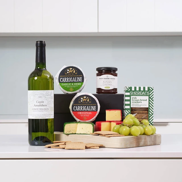 White Wine & Cheese Gift Box. A wooden serving board holds various cheeses, grapes, and crackers. Behind it are two rounds of Carrigaline cheese, a jar of chutney, and a box of Sheridan's Crackers. A bottle of white wine stands beside the food arrangement on a kitchen countertop.