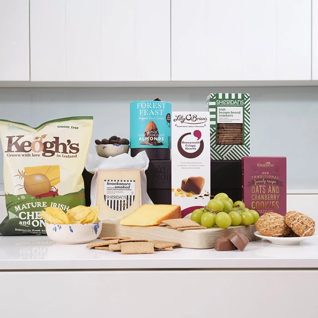 Gourmet Delights Hamper. A selection of gourmet snacks is displayed on a light-colored countertop. Items include a bag of Keogh's cheese and onion crisps, various cheeses, crackers, grapes, chocolate bars, and cookies. Several product boxes are arranged in the background, showcasing brands like Forest Feast and Lily O'Brien’s. The overall presentation suggests a curated food assortment ideal for entertaining or snacking.