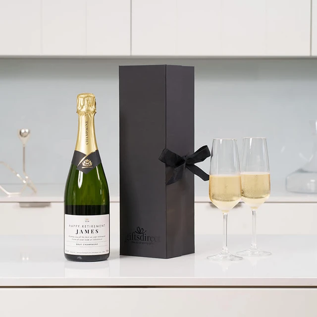 Personalised retirement champagne gift featuring a custom-labeled bottle reading ‘Happy Retirement James,’ displayed with two filled flutes and a black ribboned gift box on a white surface.