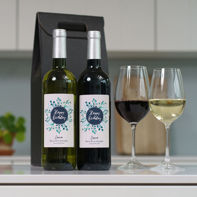 Happy Birthday Personalised Wine Duo featuring custom-labeled red and white wine bottles, paired with filled wine glasses and a black gift bag in a kitchen setting.