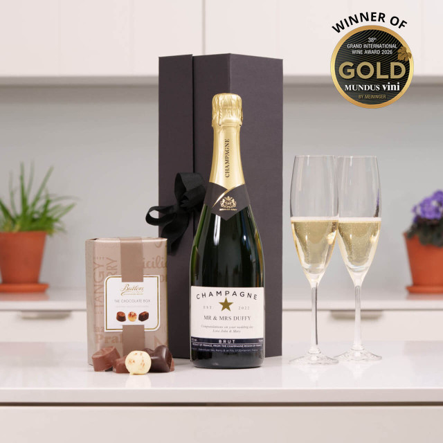 Personalised Champagne with Galway Flutes & Chocolates: A bottle of champagne is displayed next to two crystal flutes and a box of chocolates. The champagne is in a black gift box with a ribbon. In the background, there are potted plants on a countertop and award winning wine badge.
