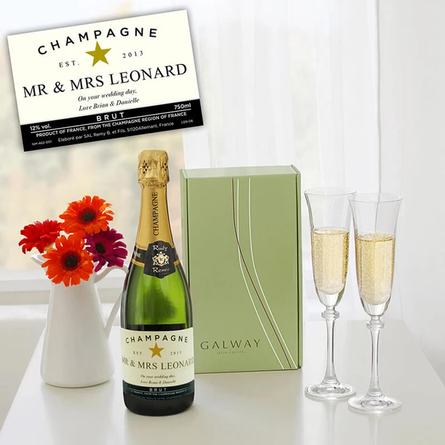 Personalised Champagne & Galway Crystal Flutes. A bottle of champagne labeled "Mr & Mrs Leonard" stands beside two filled champagne flutes. The background features a light-colored window with a bouquet of red flowers in a white vase. A green gift box, partially visible, accompanies the champagne.