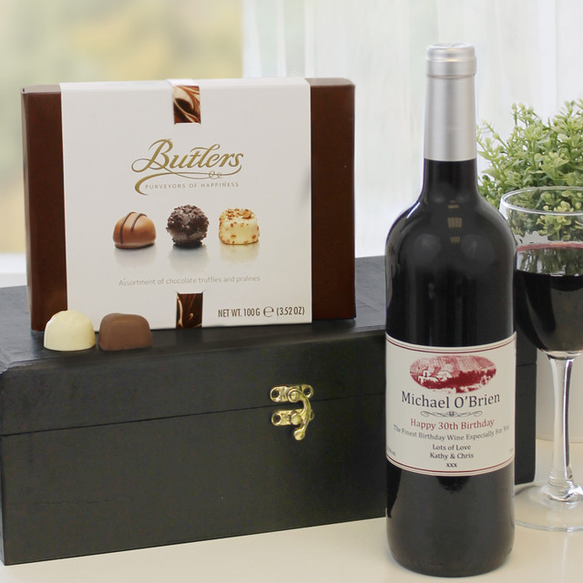 Personalised Red Wine and Chocolates: A box of Butlers chocolates sits beside a bottle of red wine labeled "Michael O'Brien." There is also a glass of the same wine on the table, with a black wooden box in the background.