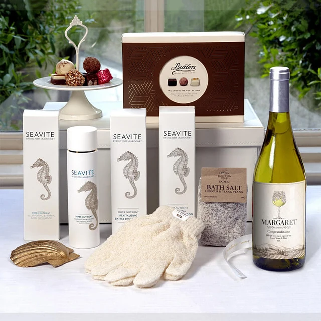 Ultimate Relaxation with Personalised Wine Pamper Gift. A flat lay arrangement featuring a selection of spa and relaxation items. The image includes two bottles of Seavite skincare products, a package of bath salt, soft gloves, and a bottle of white wine personalised with the name Margaret. In the background, there is an elegant box of chocolates on a pedestal, along with decorative truffles. The setting has natural light and greenery visible through the window.