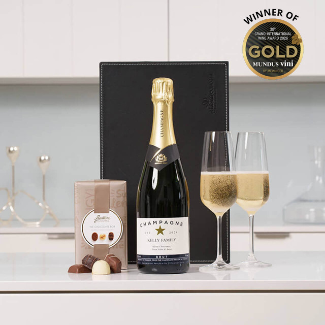 Personalised Champagne & Chocolates: A bottle of champagne sits on a white countertop with two fluted glasses filled partially. Next to the champagne is a Butler's box of chocolates, and a sleek black box is visible in the background.